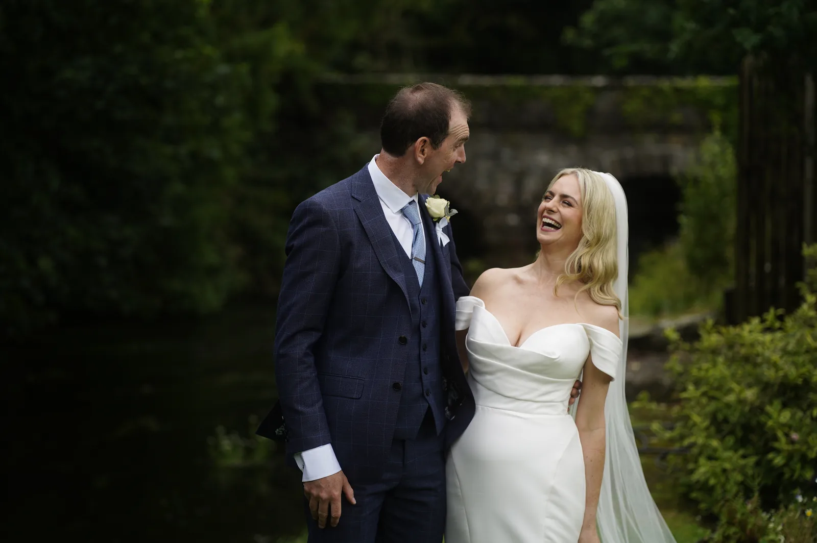 Bride and groom laughing together in a garden, groom in a blue suit