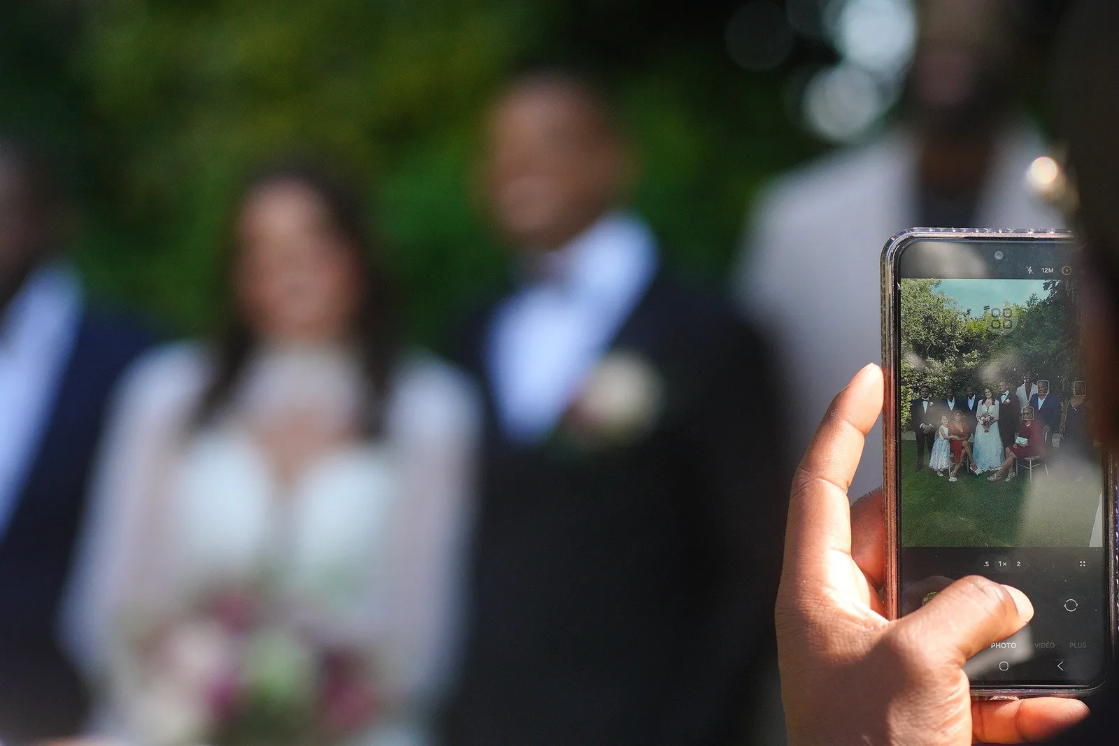 A wedding guest filming the couple on a phone, with photo and video modes visible on screen